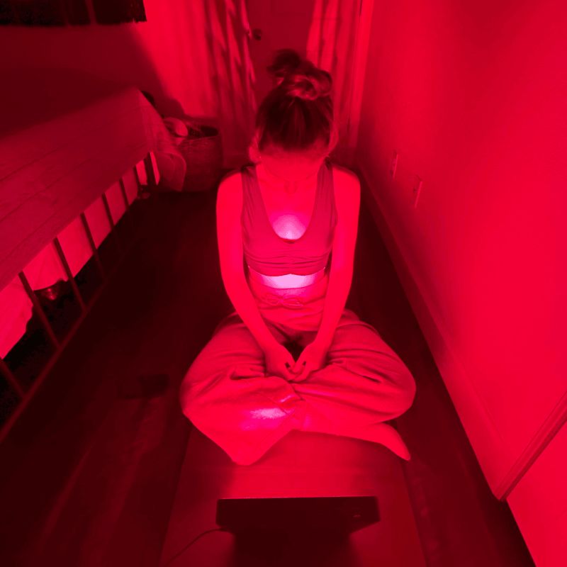 Person sitting on a chair with a Fringe Red Light Panel on their back in a dimly lit room.